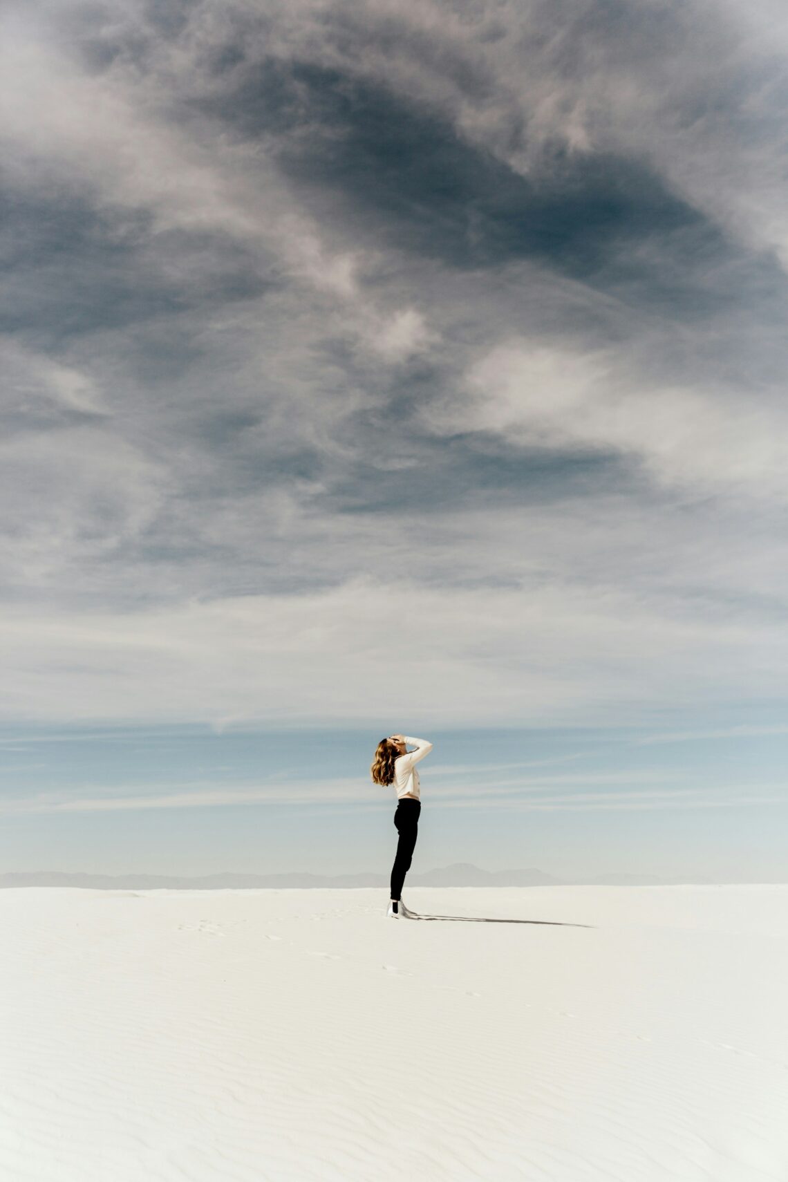Lady standing on a beach with white sand with her hands on her face looking up to the sky of grey clouds over head with the clear calm ocean