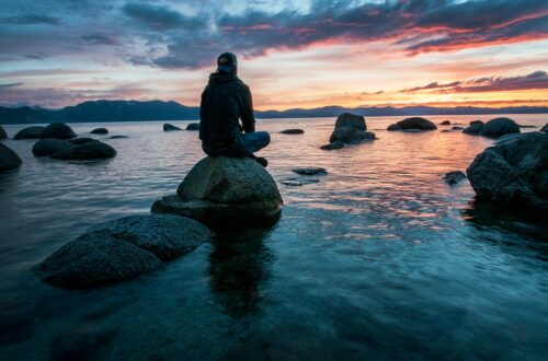 Man sitting on a rock surrounded by what deep in thought with a somber emotion watching the sun set with dim light
