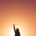 Woman with her hand in the air celebrating an achievement strength and determination with orange sky and sun behind her